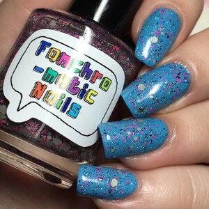 以下が含まれることがあります： 「Fonchromatic Nails」と書かれたラベルが付いたマニキュアのボトル。ボトル内のマニキュアは、ピンク、シルバー、ブラックのグリッターのミックスです。爪は、ピンク、シルバー、ブラックのグリッターが入った明るいブルーに塗られています。