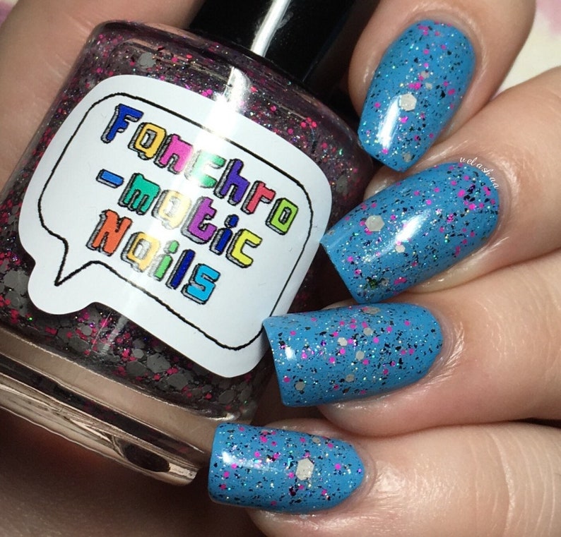 以下が含まれることがあります： 「Fonchromatic Nails」と書かれたラベルが付いたマニキュアのボトル。ボトル内のマニキュアは、ピンク、シルバー、ブラックのグリッターのミックスです。爪は、ピンク、シルバー、ブラックのグリッターが入った明るいブルーに塗られています。