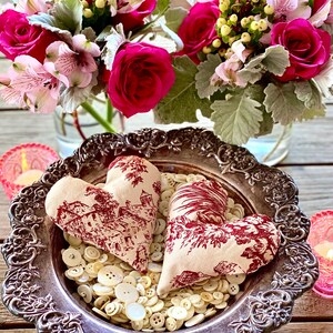 May include: Two fabric hearts with a red and white toile pattern are nestled in a silver bowl filled with white buttons. The bowl is on a wooden table with two pink glass candle holders.