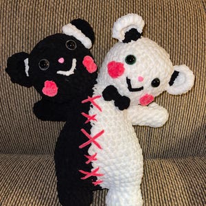 May include: Two crocheted plush toys sewn together. One is black with white eyes and a pink blush, the other is white with green eyes and a pink blush. They are both smiling and have pink bows. The toys are stitched together with pink thread.