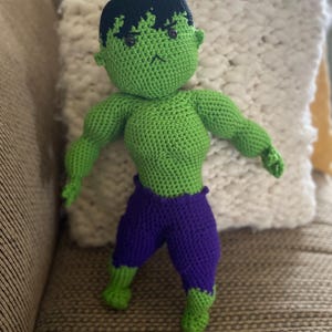May include: A green crocheted Hulk doll with a black hairpiece, purple pants, and a grumpy expression.
