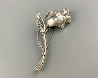 Könnte beinhalten: Eine silberne Rosenbrosche mit einem detaillierten Blattdesign. Die Rose ist realistisch gestaltet und der Stiel ist aus gedrehtem Draht.