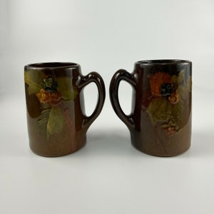 May include: Two brown ceramic mugs with floral designs. The mugs have brown, green, orange, and black painted details.