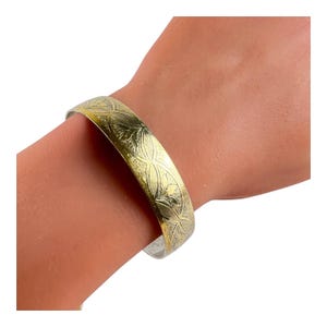 May include: A gold-colored bangle bracelet with an engraved floral pattern. The bracelet is worn on a wrist and has a simple, elegant design. The metal has a slightly aged appearance.