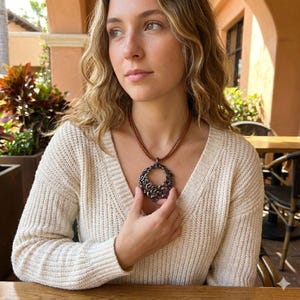 Puede incluir: Una mujer lleva un collar de cuero marrón con un colgante grande y ornamentado de metal oscuro. El colgante tiene un diseño circular con intrincados patrones florales. La mujer lleva un suéter color crema.