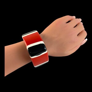May include: A bold, rectangular bracelet in red, black, and white. The bracelet features a wide, glossy red band with a central black section and white accents. The bracelet is worn on a wrist against a black background.