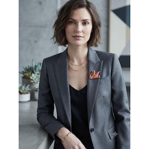 May include: A copper-toned swan brooch pinned to the lapel of a charcoal gray blazer. The blazer is worn over a black top, complemented by a delicate gold-toned chain necklace and a matching link bracelet on the wrist, creating a polished look.