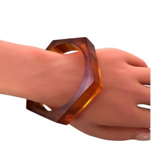 May include: A hexagonal bangle bracelet crafted from translucent amber-colored resin with deep brown marbled swirls. The rigid, geometric accessory features a smooth, polished finish and a wide band design that encircles the wrist.