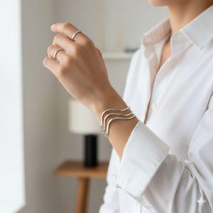 Könnte beinhalten: Ein silbernes Armband mit wellenförmigem Design und zwei passenden silbernen Ringen. Der Schmuck wird an der Hand einer Person getragen, mit einem weißen Hemd im Hintergrund. Das Armband hat drei parallele Wellenlinien.