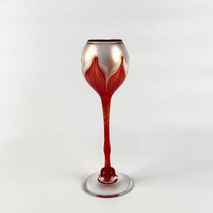 May include: A red and white glass goblet with a long stem and a round base. The goblet has a white iridescent finish and a red and white floral design.