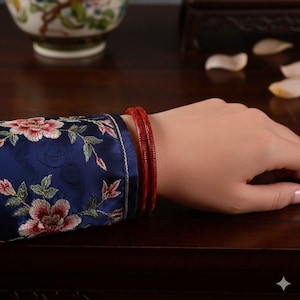 Puede incluir: Dos pulseras rojas con un diseño tallado en una muñeca. La persona lleva una manga azul oscuro con bordados florales en rojo, rosa y verde. El fondo es una superficie de madera oscura.
