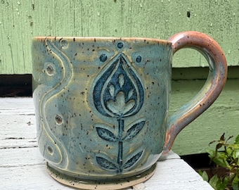 May include: A ceramic mug with a speckled teal and blue glaze featuring a carved floral motif on the front and a wavy line pattern on the side. The mug has a sturdy, curved handle with a reddish-brown gradient finish and a wide, circular base.