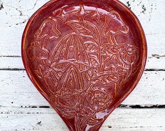 May include: A teardrop-shaped ceramic spoon rest in a deep glossy red glaze. The surface features an embossed design of mushrooms, leaves, and small four-pointed stars, with the raised lines creating a textured pattern across the entire dish.