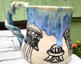 May include: A ceramic mug featuring a twisted handle and a cream-colored base decorated with black ink illustrations of jellyfish. The upper portion of the mug is glazed in a dripping blue and light green finish that cascades over the jellyfish.