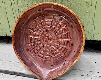 May include: A round ceramic spoon rest with a glossy, mottled reddish-brown glaze. The interior features a raised, textured spiderweb pattern radiating from the center. The rim is slightly uneven, and a small hole is visible at the top edge.