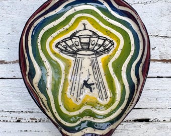 May include: A ceramic spoon rest featuring a hand-drawn black ink illustration of a UFO beaming up a person. The background consists of concentric, wavy bands of glossy yellow, green, blue, and deep red glaze with a speckled, off-white center.