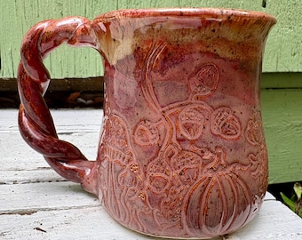May include: A ceramic mug with a glossy, variegated red and brown glaze. The exterior features embossed designs of pumpkins, acorns, and mushrooms. The handle is shaped like a twisted rope, and the rim has a smooth, slightly flared edge.