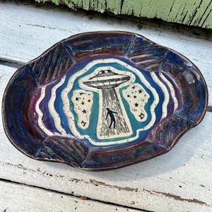May include: An oval ceramic dish featuring a central illustration of a UFO beaming down a silhouette of a person. The design includes blue and purple wavy borders with star accents. The glaze is a deep, glossy mix of dark blue and purple tones.
