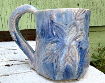 May include: A ceramic mug with a glossy finish in shades of blue and white. The surface features a hand-carved floral pattern with embossed petals and leaves. It has a thick, curved handle and a slightly irregular, organic shape throughout the body.