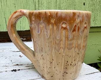 May include: A ceramic mug with a glossy finish featuring a tan base with dark brown speckles and a darker brown drip glaze pattern cascading from the rim. The mug has a thick, curved handle and a smooth, rounded body with subtle vertical indentations.