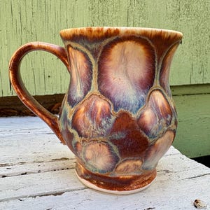 May include: A ceramic mug featuring a glossy glaze with a repeating pattern of circular shapes in shades of deep brown, tan, and muted blue. The mug has a curved body, a wide rim, and a single handle attached to the side of the vessel.