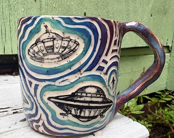 May include: A ceramic mug featuring a hand-carved design of black flying saucers and a small silhouette of a person against a background of swirling blue, teal, and purple glaze. The mug has a speckled finish and a curved, glazed handle.