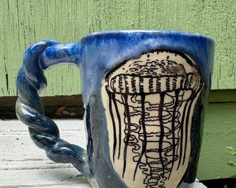 May include: A ceramic mug featuring a blue and white glazed finish with a prominent black line drawing of a jellyfish on the front. The mug has a distinct twisted, rope-like handle in a deep blue glaze that contrasts with the lighter body.