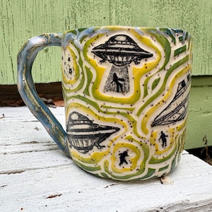 May include: A ceramic mug featuring a hand-carved relief pattern of black UFOs and silhouettes of figures against a background of swirling yellow and green glaze. The handle has a marbled blue and grey finish with a speckled texture throughout.