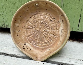 May include: A round, beige ceramic grater plate with a textured, radial grid pattern in the center for grating. The surface features dark brown speckles throughout the glaze, and the dish includes a small, pointed spout at the bottom edge.