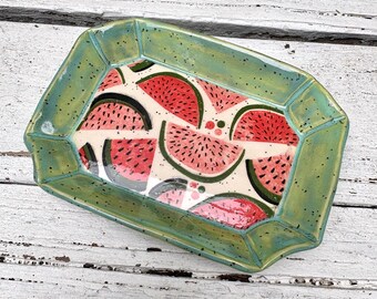 May include: A rectangular ceramic tray with a glossy, speckled light green rim and a white interior base featuring a repeating pattern of red watermelon slices with black seeds and dark green rinds, arranged in a scattered, overlapping design.