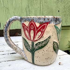 May include: A ceramic mug with a cream-colored base featuring a speckled texture and a blue-toned rim. The exterior is decorated with hand-carved floral motifs, including a prominent red tulip with green leaves and a partial green flower design.