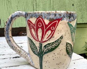 May include: A ceramic mug with a cream-colored base featuring a speckled texture and a blue-toned rim. The exterior is decorated with hand-carved floral motifs, including a prominent red tulip with green leaves and a partial green flower design.