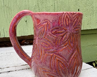 May include: A ceramic mug with a glossy magenta and deep pink glaze featuring a carved, repeating floral petal pattern across the exterior surface. The mug has a curved handle on the left side and a smooth, rounded body with a consistent color finish.