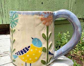 May include: A ceramic mug with a cream-colored base featuring a hand-painted yellow bird with blue polka dots and a blue head. The mug is decorated with embossed green stems, a teal flower, and an orange flower. It has a thick, glossy blue handle.