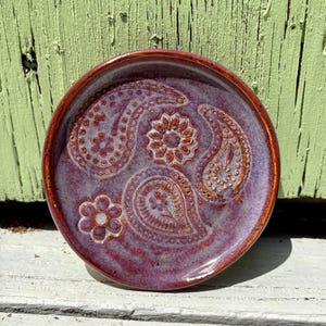 May include: A round ceramic dish featuring a glossy purple and reddish-brown glaze. The surface is decorated with embossed paisley patterns and small flower motifs, with a raised rim that matches the reddish-brown color of the glaze accents.