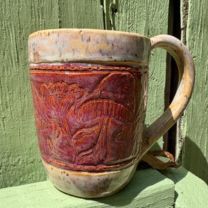 May include: A ceramic mug featuring a deep red central band with embossed floral and mushroom designs. The top and bottom sections of the mug are finished in a speckled beige glaze. It includes a curved handle on the right side and a smooth rim.
