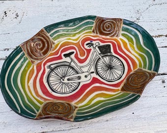 May include: An oval ceramic dish featuring a central black line drawing of a bicycle with a basket. The background consists of wavy, concentric bands of green, yellow, orange, and red. Four raised, brown, spiral-textured squares accent the rim.