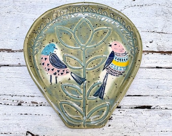 May include: A teardrop-shaped ceramic spoon rest with a speckled sage green glaze. The surface features a central embossed floral stem flanked by two birds, one with a blue head and pink body, the other with a pink head and yellow-banded body.