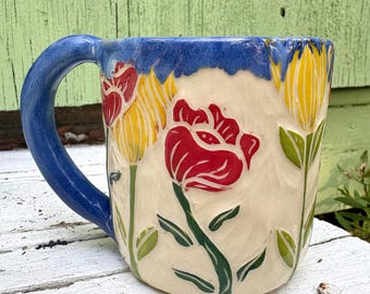 May include: A ceramic mug with a cream-colored base featuring carved and painted floral designs. The mug has a blue handle and a blue rim with a drip-like pattern. The flowers are depicted in red and yellow with green stems and leaves.