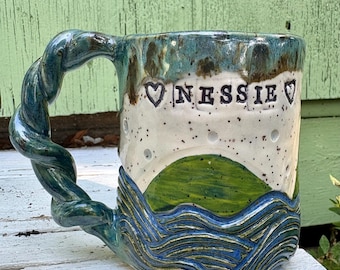 May include: A ceramic mug with a twisted handle, featuring a white upper section with the text NESSIE flanked by two hearts, a green hill, and a blue base with textured wave patterns. The glaze transitions from dark blue to white with speckles.