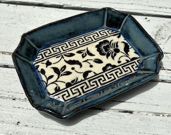 May include: A rectangular ceramic tray with a dark blue rim and a cream-colored center. The center features a black floral and geometric pattern. The tray has a glossy finish and is set against a white wooden surface.