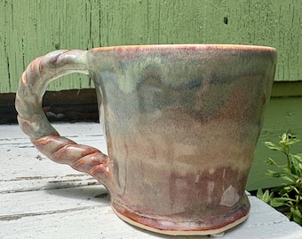 May include: A ceramic mug featuring a multi-colored glaze with blended tones of sage green, muted blue, and earthy brown. The mug has a distinct, thick handle shaped like a twisted rope, and the overall surface has a smooth, glossy finish.
