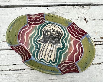 May include: An oval ceramic tray featuring a central black and white line drawing of a jellyfish. The tray has a speckled olive green border and alternating sections of red and green wavy lines surrounding the central jellyfish illustration.