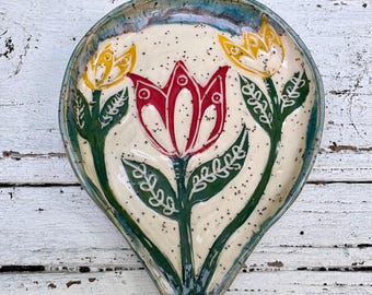 May include: A teardrop-shaped ceramic spoon rest with a cream-colored base featuring dark speckles. The surface displays a hand-painted design of a large central red tulip flanked by two smaller yellow tulips, all with green stems and leaves.