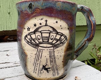May include: A ceramic mug with a reactive glaze in shades of purple, blue, and brown. The front features a black ink illustration of a flying saucer beaming down a small figure, surrounded by stars, set against a cream-colored circular background.