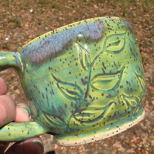 May include: Handmade ceramic mug with a green and blue glaze. The mug features an embossed leaf design and a speckled, off-white base. The handle is integrated into the design, and the mug is held by a hand.