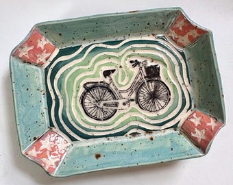 May include: Rectangular ceramic tray with a teal border and a bicycle illustration. The center features a bicycle design with green and white wavy lines. The corners have red and white star accents. A decorative piece for the home.