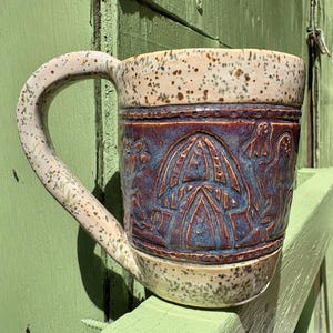 May include: A ceramic mug with a speckled cream-colored glaze on the top and bottom sections. The center band features a deep purple glaze with embossed, carved mushroom designs. The mug includes a large, matching speckled handle on the side.