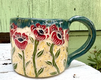 May include: A ceramic mug featuring a hand-painted design of red poppy flowers with green stems and leaves against a speckled cream-colored background. The rim and the handle are finished in a glossy dark teal glaze, creating a contrast.