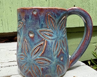 May include: A ceramic mug featuring a glossy glaze in shades of deep blue, purple, and reddish-brown. The exterior surface is decorated with embossed floral patterns and small circular indentations, complemented by a curved handle on the right side.